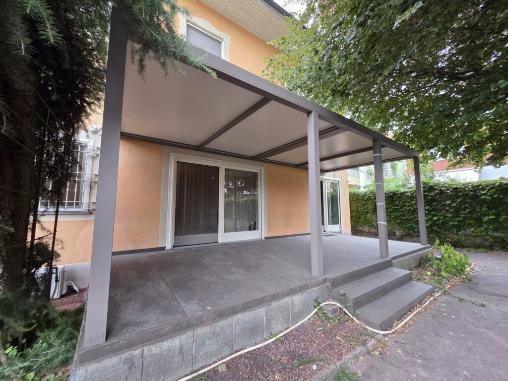 Modern outdoor patio with a gray metal frame canopy over a sliding glass door. It's attached to an orange stucco house with steps leading to a concrete terrace, surrounded by trees.